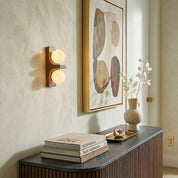 Walnut Eveline double wall sconce short version beside gold framed abstract artwork above dark console with books and vase
