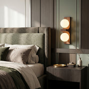 Walnut Eveline double wall sconce short version glowing above dark wood nightstand beside upholstered bed headboard