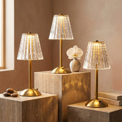 Three golden crystal table lamps in different sizes displayed on travertine pedestals with orchid vase