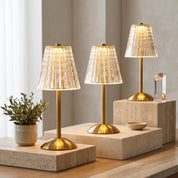 Three golden crystal table lamps glowing on travertine blocks with green plant in minimal setting