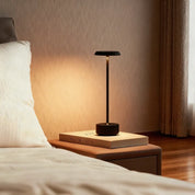 Black Luxora cordless table lamp glowing warm on bedside bench beside book in neutral bedroom