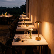 Gold Luxora cordless table lamp as centerpiece on round outdoor terrace dining table with wine glasses under string lights at night