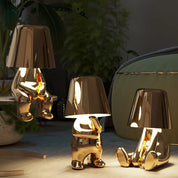 Three gold RayDudes sculptural table lamps in different poses glowing in dark room with plants